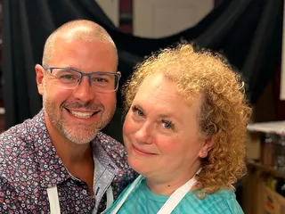 Andy and his wife Alicia, arms around each other and both smiling, in aprons.