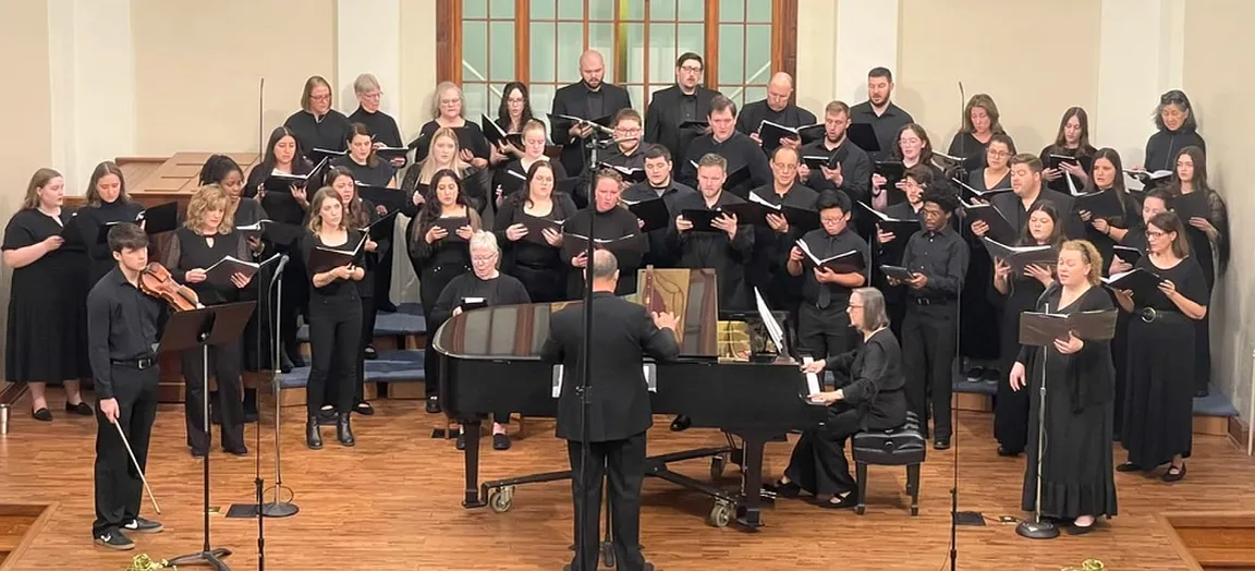 The choir on stage in concert dress, singing from black folders, with a pianist, a violinist and a soloist, conducted from the front.