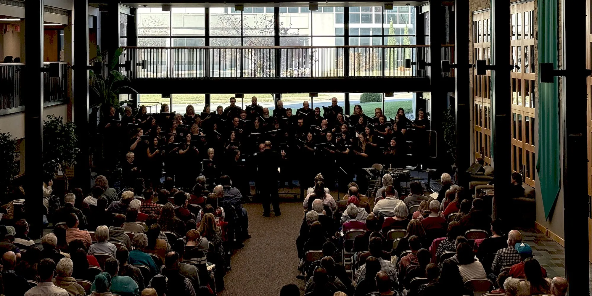 The choir in concert dress performing to a full audience in a glass-walled atrium.