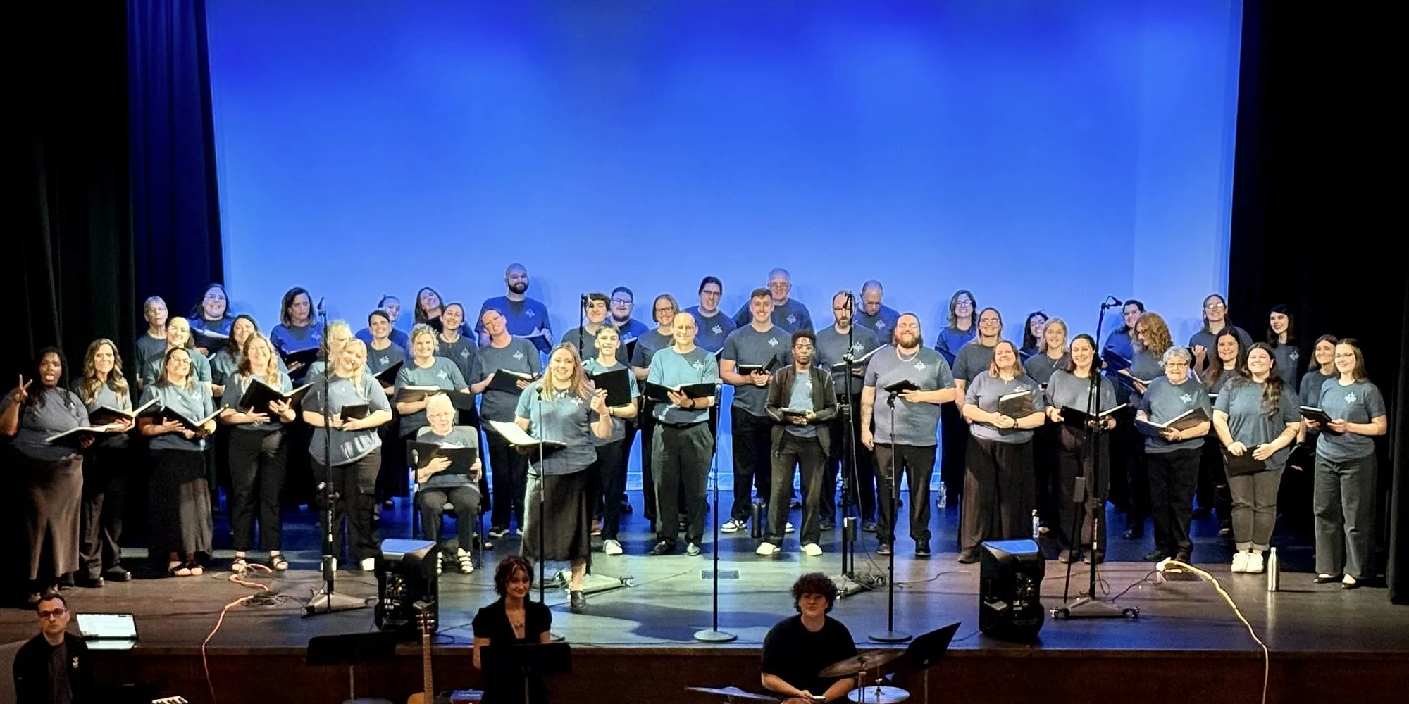 The choir on stage in matching shirts, warming up before a concert with the band below them.