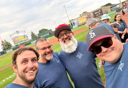 Four choir members in matching choir t-shirts, grinning, on the grass at a baseball stadium before singing.