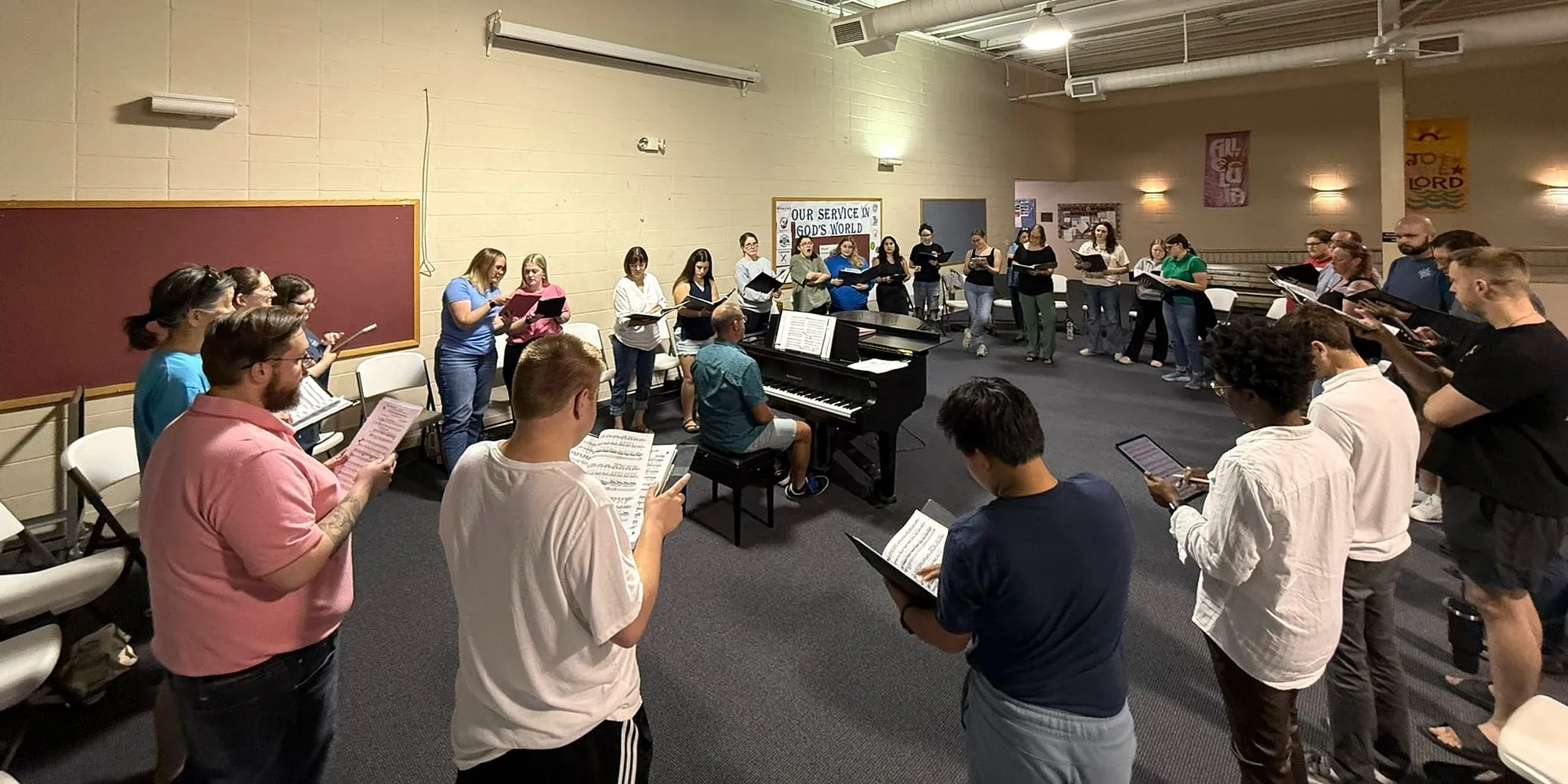 Singers in everyday clothes standing in a circle around a piano at rehearsal, folders open.