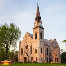 Stone Chapel at Drury University at dusk — a stone church with a tall copper spire and a lit clock tower, floodlit against a pale evening sky.