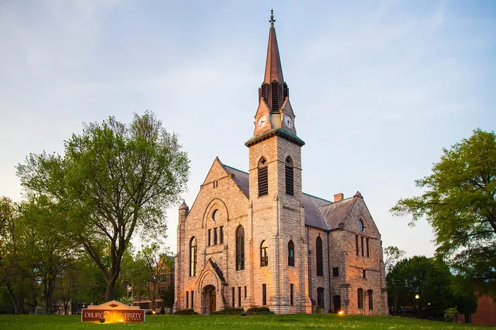 Stone Chapel at Drury University at dusk — a stone church with a tall copper spire and a lit clock tower, floodlit against a pale evening sky.